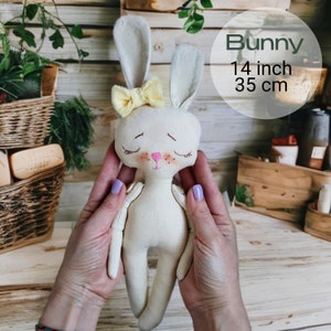 May include: A white fabric bunny doll with a yellow bow, pink cheeks, and closed eyes. The doll is 14 inches tall and is being held by a person's hands.
