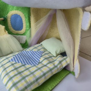 May include: A handmade felt dollhouse interior with a bed, pillow, and window. The bed has a green, yellow, and white plaid pattern with a blue and white patch. The pillow is light green, and the window has a blue center.