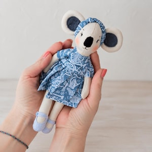 May include: A small fabric koala doll with cream-colored skin, large round ears, and pink cheeks. The doll wears a blue dress featuring a white bicycle and floral pattern, a matching blue headband, and light blue shoes with a single strap closure.