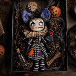 May include: A handcrafted doll with large purple bat ears, a stitched mouth, and a ruffled collar. The doll wears a grey striped dress with orange and purple patches, black sleeves, and suspenders. It is displayed in a box with a small pumpkin, a glass bottle, a miniature broom, and a key.