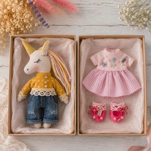 May include: Two gift boxes with a handmade unicorn doll and a set of doll clothes. The unicorn has a yellow horn, a yellow and white mane, and is wearing a yellow shirt and denim jeans. The doll clothes include a pink dress and pink shoes.