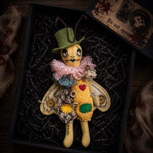 May include: A handmade fabric doll with a yellow face, stitched features, and a green top hat. It wears a pink lace ruff, a yellow polka-dot body with a red heart patch, floral fabric accents, and two large wings. A box lid nearby reads Do not open.