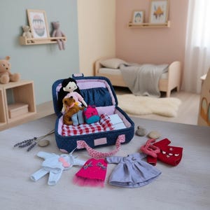 Può includere: Una valigia blu a pois aperta rivela una bambola, orsacchiotti e un piccolo zaino. Diversi completi per bambole, tra cui un vestito rosa con tulle, un vestito azzurro e un completo rosso, sono disposti nelle vicinanze. Tema: casa delle bambole o gioco per bambini.