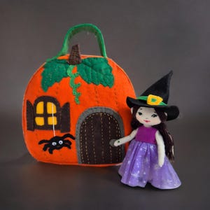 Op de afbeelding: Een vilten Halloween-decoratie met een oranje pompoenvormig huis met een groene handgreep en bladeren. Een zwarte spin staat bij de deur. Een pop verkleed als heks in een paarse jurk en zwarte hoed staat naast het huis.