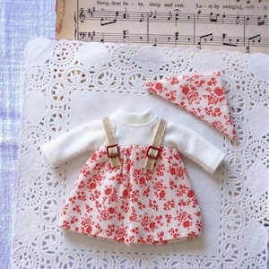 May include: A miniature doll outfit featuring a white long-sleeved top and a red floral print dress with tan straps. A matching red floral print bandana is also included. The outfit is displayed on a white lace doily.