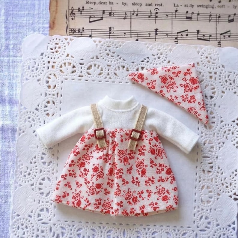 May include: A miniature doll outfit featuring a white long-sleeved top and a red floral print dress with tan straps. A matching red floral print bandana is also included. The outfit is displayed on a white lace doily.