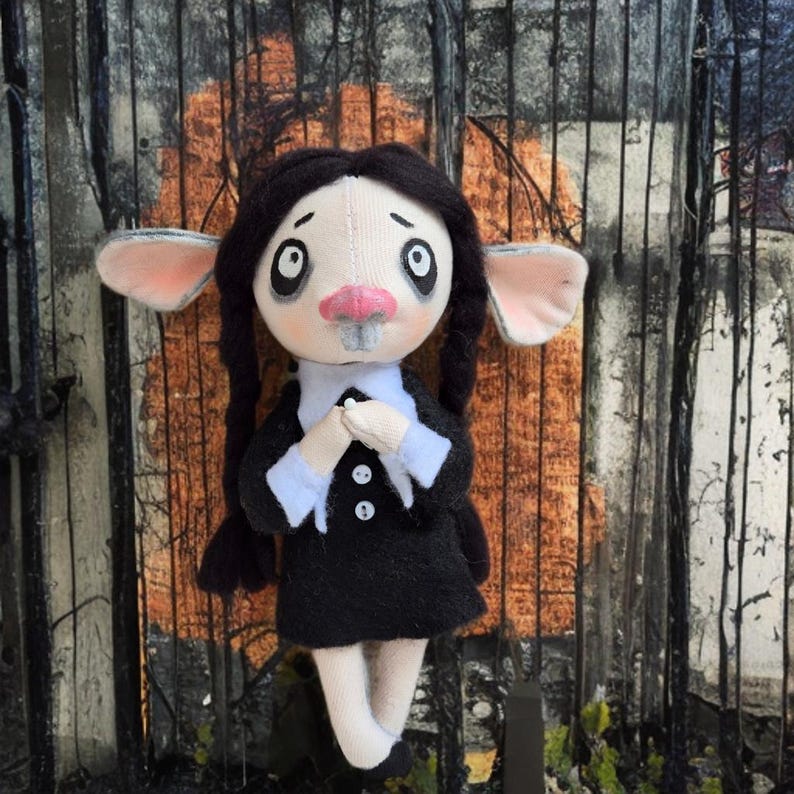 May include: A handmade fabric doll with a pale face, large dark eyes, and a pink nose. The doll has long black braided hair, large ears, and wears a black dress with a white collar and cuffs, featuring two small white buttons on the front.