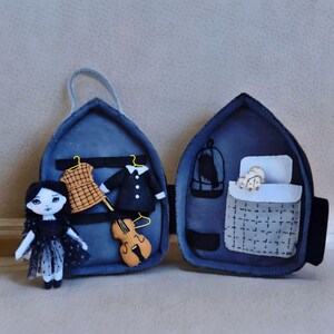 Op de afbeelding: Een handgemaakte vilten pop in een zwarte jurk staat naast een open blauwe vilten koffer. Binnenin bevinden zich miniatuurkleding, een viool, een vogelkooi en een bed met een slapende kat. De koffer heeft een handvat.