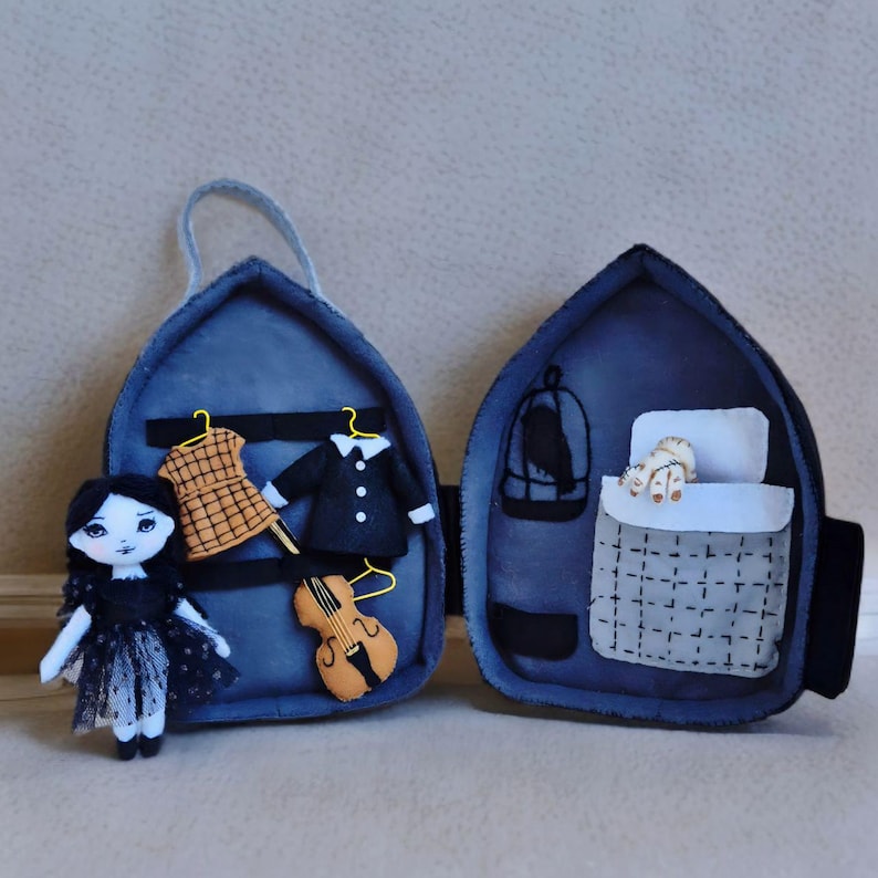 Op de afbeelding: Een handgemaakte vilten pop in een zwarte jurk staat naast een open blauwe vilten koffer. Binnenin bevinden zich miniatuurkleding, een viool, een vogelkooi en een bed met een slapende kat. De koffer heeft een handvat.