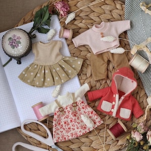 May include: A collection of miniature doll clothes, including a tan dress with white polka dots, a pink sweater, brown pants, a red hooded jacket, and a floral print dress. Accessories include spools of thread and a small embroidered container.