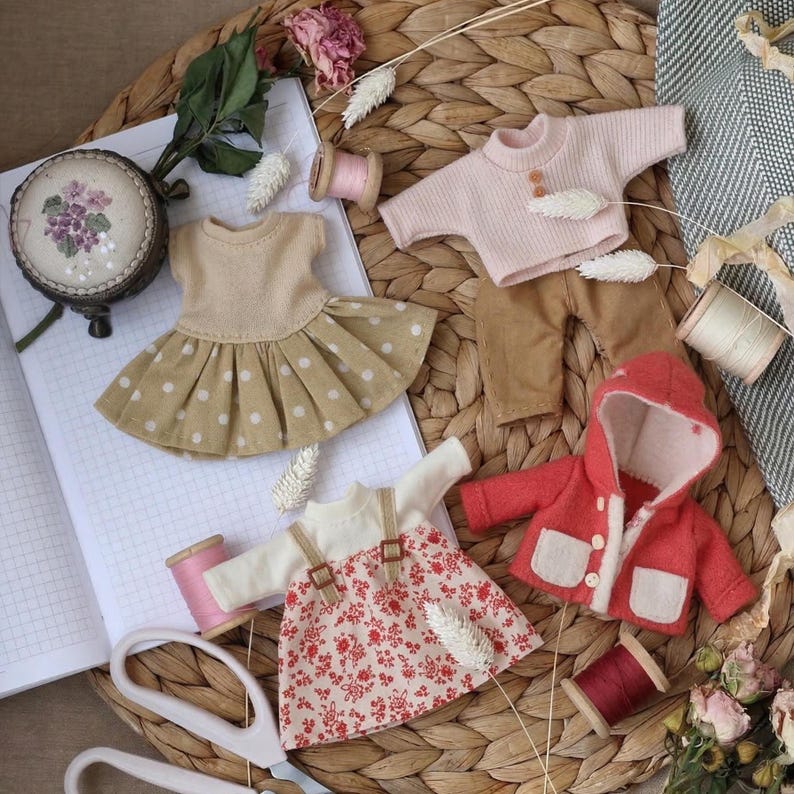 May include: A collection of miniature doll clothes, including a tan dress with white polka dots, a pink sweater, brown pants, a red hooded jacket, and a floral print dress. Accessories include spools of thread and a small embroidered container.