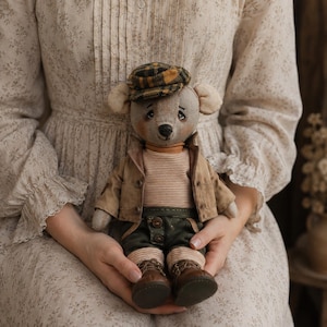 May include: A stuffed bear toy wearing a yellow and black plaid flat cap, a tan jacket, a beige and white striped shirt, dark green trousers with buttons, and brown boots. The bear has light brown fur, stitched facial features, and round ears.
