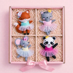 May include: A set of four felt animal dolls in a pink box. The dolls include a fox, cat, mouse, and panda, each wearing a tutu and flower crown. The box is tied with a pink ribbon.