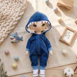 Puede incluir: Muñeca hecha a mano con un disfraz de tiburón de pana azul con capucha y dientes de fieltro blanco. La muñeca tiene pelo rubio rizado, ojos verdes y zapatillas blancas. Un pequeño juguete de tiburón está al lado de la muñeca.