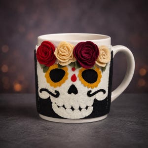 May include: A white ceramic mug featuring a felt wrap with a sugar skull design. The skull has black eye sockets with yellow scalloped borders, a red teardrop detail, and a black jaw. A row of red and cream felt roses sits above the skull.