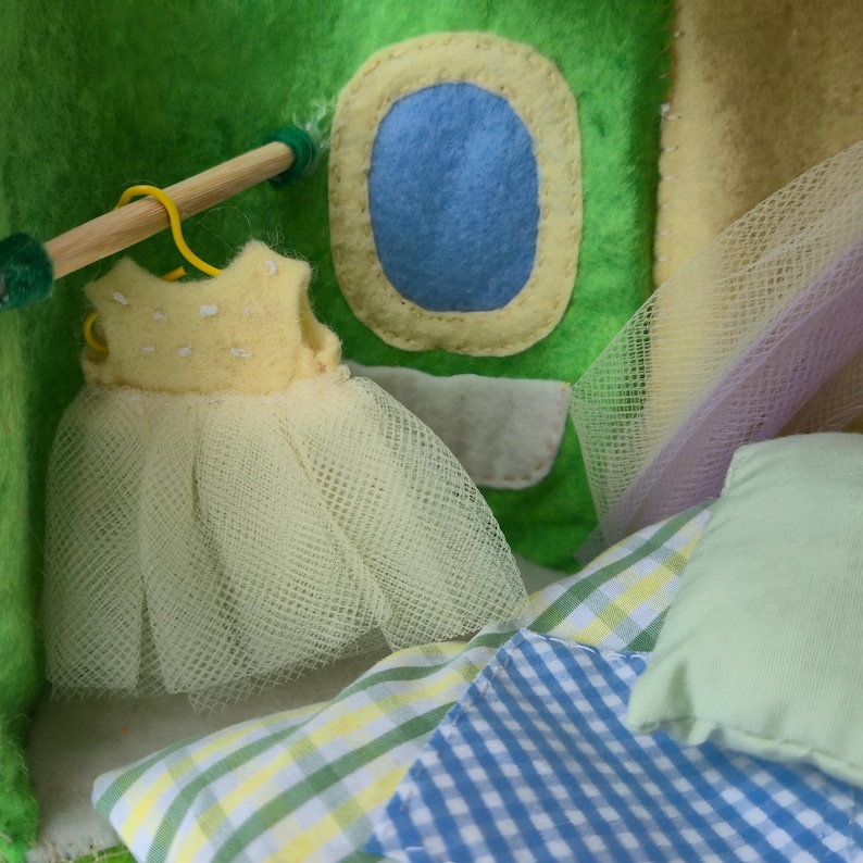 May include: A miniature dollhouse scene with a yellow dress on a hanger, a blue window, and a patchwork quilt. The dollhouse is made of green and yellow felt. A wooden dowel with green ends serves as a clothes rod.