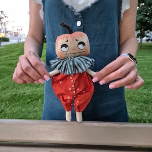 Puede incluir: Una mu&ntilde;eca de calabaza hecha a mano con un dise&ntilde;o caprichoso. La mu&ntilde;eca presenta una cabeza en forma de calabaza con rasgos faciales cosidos, un cuello a rayas y un mono rojo con patrones de estrellas blancas. La mu&ntilde;eca se sostiene sobre un fondo verde.