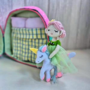 Pu&ograve; includere: Una bambola di feltro fatta a mano con capelli rosa e un vestito verde che cavalca un unicorno blu con un corno giallo. La bambola &egrave; di fronte a uno zaino di feltro rosa e verde con un letto all'interno.