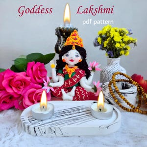 May include: A handcrafted Goddess Lakshmi doll, adorned with a golden crown and jewelry, sits on a decorative tray with lit candles. The image includes pink roses, a vase of yellow flowers, and the text "Goddess Lakshmi pdf pattern."