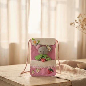 May include: Pink felt crossbody bag with a grey mouse character wearing a green cape and holding a strawberry. The bag features white floral accents, a white lace trim, and a pink cord strap. The bag is approximately 15 cm tall.