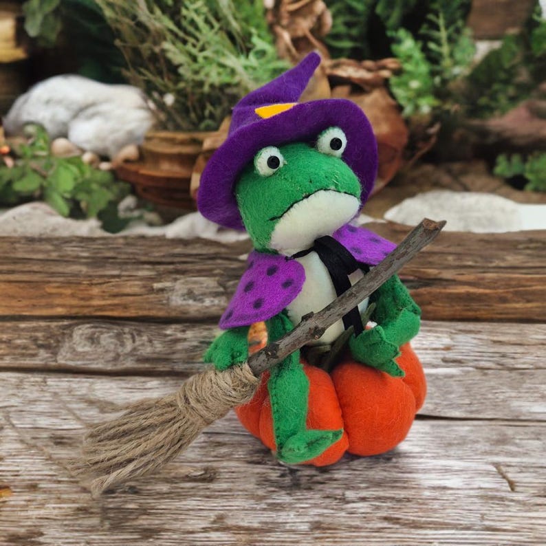 May include: A handmade felt frog wearing a purple witch hat with a yellow accent and a purple cape with polka dots. The frog is holding a twig broom and sitting on an orange pumpkin. The frog has a black tie and a white shirt.