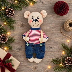 May include: A stuffed bear toy with a cream-colored fabric body, pink cheeks, and a hand-drawn black nose and mouth. The bear wears a red and white horizontal striped short-sleeve shirt paired with blue denim overalls featuring two green buttons.