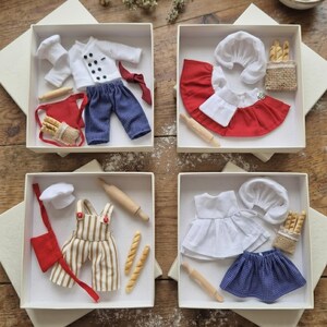 Puede incluir: Cuatro juegos en caja de trajes de panadero en miniatura. Cada juego incluye un gorro de chef, un delantal y ropa. Un juego tiene una chaqueta de chef blanca y pantalones a cuadros azules. Otro tiene un atuendo rojo y blanco. El tercero tiene un mono a rayas beige y blanco. El cuarto tiene un vestido blanco y una falda a cuadros azules.