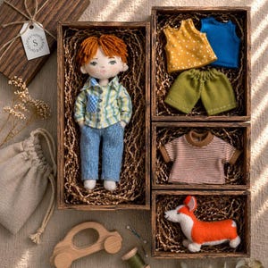 May include: A handmade doll with red hair, wearing a plaid shirt and jeans, is displayed in a wooden box. Additional wooden boxes contain doll clothing and a felt corgi dog. A wooden toy car and sewing supplies are also present.