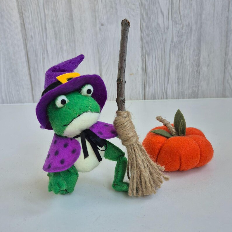 May include: A green frog plush toy wearing a purple witch hat and cape with black polka dots. The frog holds a broom made of a twig and twine. An orange felt pumpkin with a green leaf sits nearby. Halloween themed.