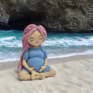 May include: A felt doll of a pregnant person with pink hair and a blue dress, seated in a meditative pose on a sandy beach. The doll's eyes are closed, and the hands rest gently on the belly. The background features a turquoise ocean and rocky cliffs.