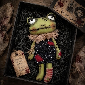 May include: A handmade green frog doll with stitched features, wearing a polka-dot outfit with red patches and a lace collar. The doll is in a black box with a tag that reads "Found on the attic. Do not wash."