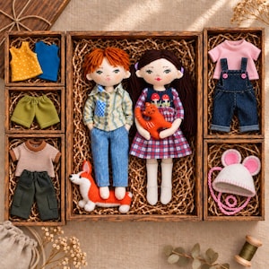 May include: Two fabric dolls with embroidered faces and yarn hair, one with ginger hair and one with dark brown hair, are presented in a wooden box with extra clothing sets, a small plush orange cat, a plush dog, and a white hat with pink ears.