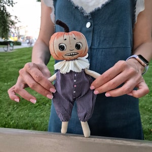 Puede incluir: Mu&ntilde;eca de calabaza hecha a mano con expresi&oacute;n preocupada. La mu&ntilde;eca tiene una cabeza en forma de calabaza con un tallo, un cuello con volantes y un mono a rayas moradas. La mu&ntilde;eca se sostiene frente a un fondo de hierba verde.