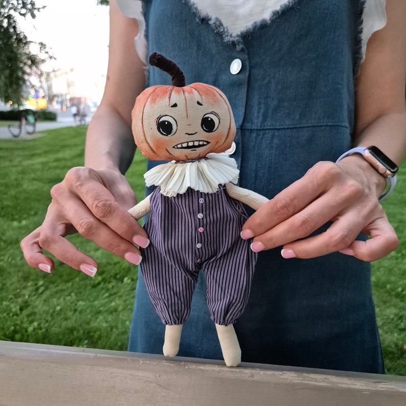 Puede incluir: Mu&ntilde;eca de calabaza hecha a mano con expresi&oacute;n preocupada. La mu&ntilde;eca tiene una cabeza en forma de calabaza con un tallo, un cuello con volantes y un mono a rayas moradas. La mu&ntilde;eca se sostiene frente a un fondo de hierba verde.