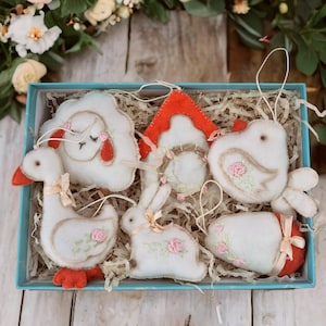 May include: A set of six felt Easter decorations in a blue box. The decorations are a white goose, a white bunny, a white bird, a white egg, a white sheep, and a red and white house. The decorations are all decorated with pink flowers.