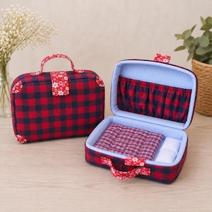 May include: Two rectangular fabric cases featuring a red and navy blue buffalo plaid pattern. One case is closed with a floral fabric handle and tab, while the other is open, revealing a light blue striped interior, a pocket, and a quilted base.
