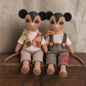 May include: Two stuffed mouse dolls with tan fabric bodies, dark brown ears, and pink noses. The left mouse wears a beige polka-dot shirt, a patchwork vest, and a green gingham scarf. The right mouse wears a striped shirt, olive overalls, and a yellow scarf.
