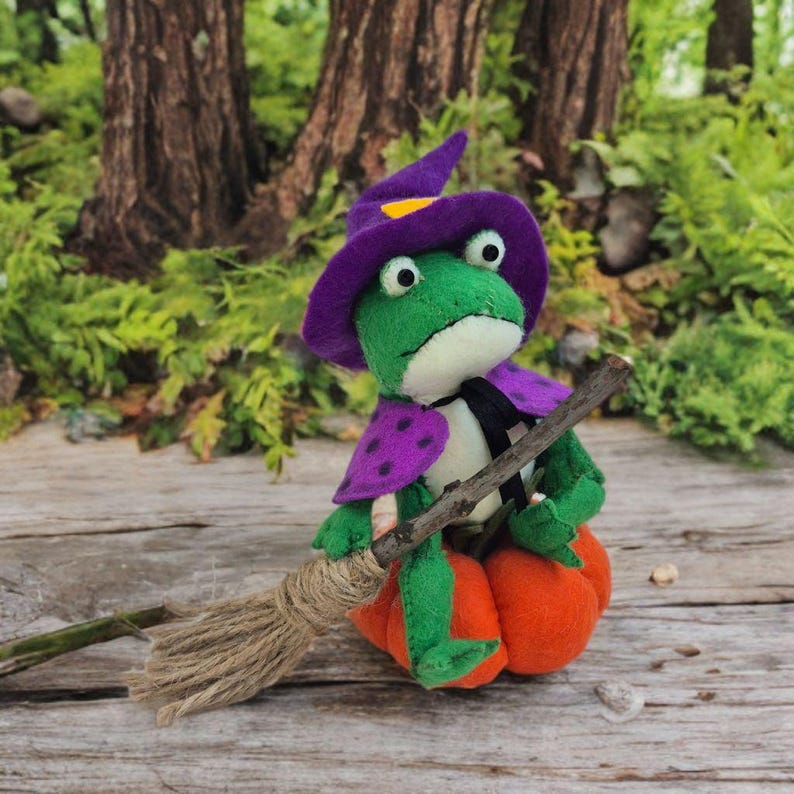 May include: A handmade green frog doll wearing a purple witch hat and cape with a black tie. The frog is sitting on an orange pumpkin and holding a broom. The background features a wooden surface and green foliage.