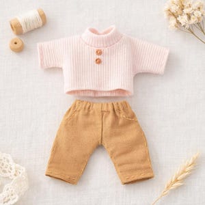 May include: A miniature doll outfit featuring a light pink ribbed knit top with two brown buttons and short sleeves. The outfit includes tan-colored pants with visible stitching details. The ensemble is set against a neutral background with craft supplies.