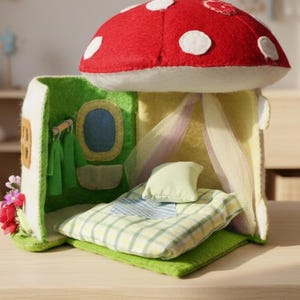 May include: A whimsical felt mushroom house with a red cap and white spots. The house features a green door, a blue window, and a bed with a plaid blanket. A small pillow and sheer curtains complete the cozy interior.