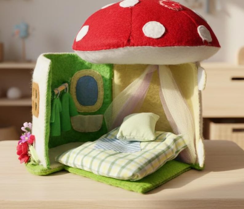 May include: A whimsical felt mushroom house with a red cap and white spots. The house features a green door, a blue window, and a bed with a plaid blanket. A small pillow and sheer curtains complete the cozy interior.
