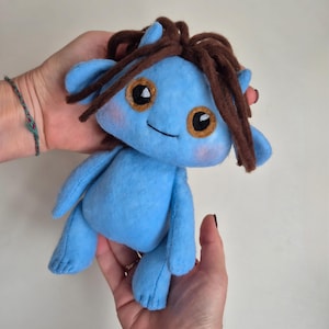 May include: A handmade blue plush doll with brown hair and large, round eyes. The doll has a smiling expression and is held in two hands. The doll's cheeks are blushed pink, and the eyes have a gold outline.