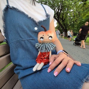 Puede incluir: Una mu&ntilde;eca de calabaza hecha a mano con una expresi&oacute;n caprichosa. La mu&ntilde;eca tiene una cabeza naranja, un cuello a rayas y pantalones rojos. La mu&ntilde;eca est&aacute; sentada en un vestido de mezclilla.