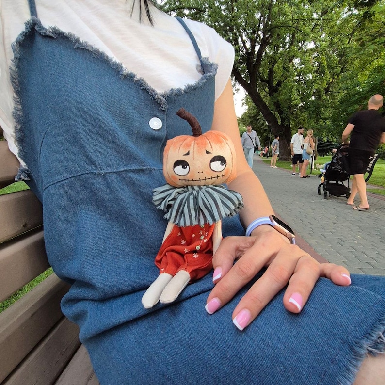 Puede incluir: Una mu&ntilde;eca de calabaza hecha a mano con una expresi&oacute;n caprichosa. La mu&ntilde;eca tiene una cabeza naranja, un cuello a rayas y pantalones rojos. La mu&ntilde;eca est&aacute; sentada en un vestido de mezclilla.