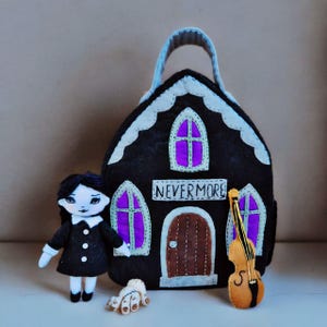 Op de afbeelding: Een zwarte vilten huisvormige tas met witte randen en het woord "NEVERMORE". Een pop in een zwarte jurk, een vilten hand en een vilten viool staan voor de tas. De ramen van de tas zijn paars.