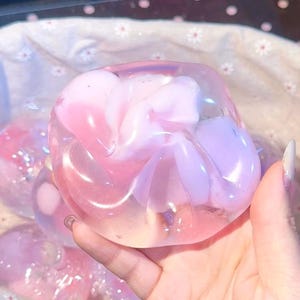 May include: A translucent, pink and lavender decorative object, possibly soap or a bath product, held in a hand. The object has a swirling, organic shape with a soft, iridescent sheen. The background is a light-coloured fabric.