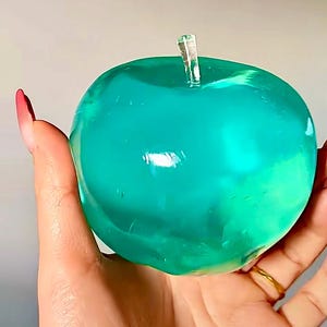 May include: A bright teal green glass apple with a clear stem. The apple is smooth and shiny, and it appears to be filled with a clear liquid.