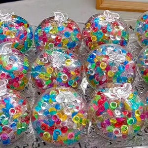 Può includere: Globi di vetro trasparente riempiti di piccole perline rotonde e colorate. Ogni globo ha una parte superiore decorativa trasparente. Le perline sono un mix di rosso, blu, verde, giallo e rosa. I globi sono disposti su una superficie riflettente.