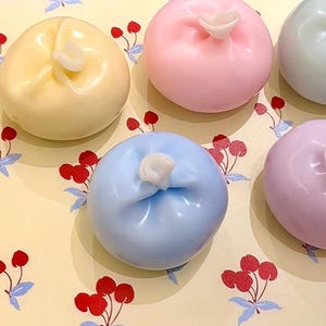 May include: Four pastel-coloured, round, fabric-covered, poufs with a gathered top, arranged on a tray with a floral pattern. The poufs are yellow, pink, blue, and purple.