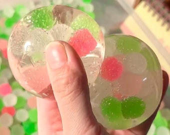 Op de afbeelding: Twee heldere, bolvormige sensorische speeltjes gevuld met kleurrijke gelparels. Eén bol wordt in een hand gehouden en toont roze, groene en witte parels. De andere bol, ook vastgehouden, bevat vergelijkbare gekleurde parels. De achtergrond is vervaagd met meer gelparels.